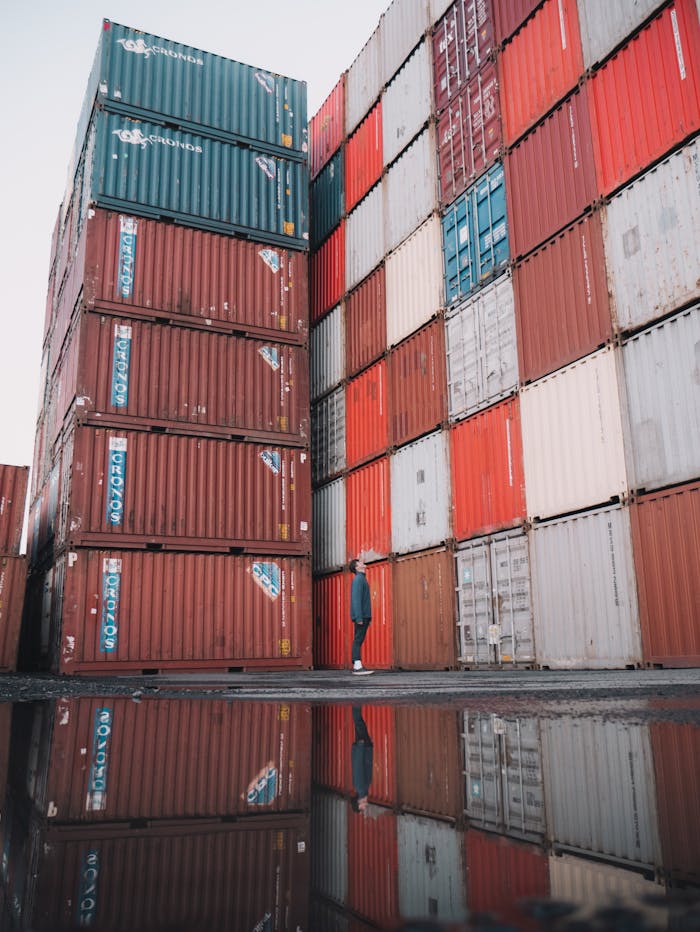 about-img A person stands beside stacked cargo containers at a port, reflecting in nearby water.